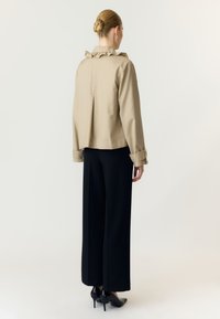 Femme avec les cheveux en chignon portant une veste beige à col à volants, un pantalon noir à jambes larges et des talons hauts noirs, debout, dos à un fond blanc.