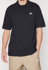 Černé bavlněné polo tričko s klasickým límcem, třemi knoflíky a bílým logem Nike na levé straně hrudníku. Volný střih, krátké rukávy.