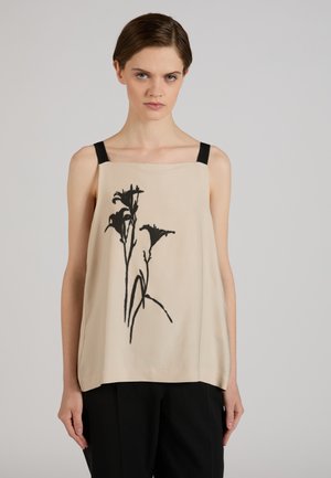 Kobieta nosząca beżowy top bez rękawów z czarnym kwiatowym wzorem i czarnymi ramiączkami, w połączeniu z czarnymi spodniami, stojąca na jednolitym tle.