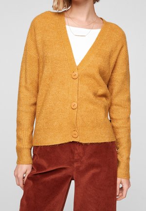 Kobieta ubrana w musztardowożółty rozpinany sweter na guziki na białej bluzce, w połączeniu z brunatno-czerwonymi sztruksowymi spodniami, prezentująca szczegóły swobodnej stylizacji.