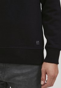Schwarzer Sweatshirt mit gerippten Bündchen und Saum. Verfügt über ein kleines Etikett mit "DNM WRKRS" auf der linken Seite nahe der Taille. Glatte Textur.