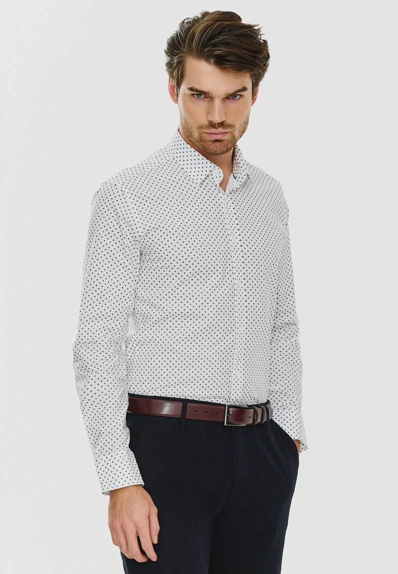 Biała koszula w małe granatowe kropki, z kołnierzykiem zapinanym na guziki, długimi rękawami i gładką teksturą, zestawiona z ciemnymi spodniami i brązowym paskiem.