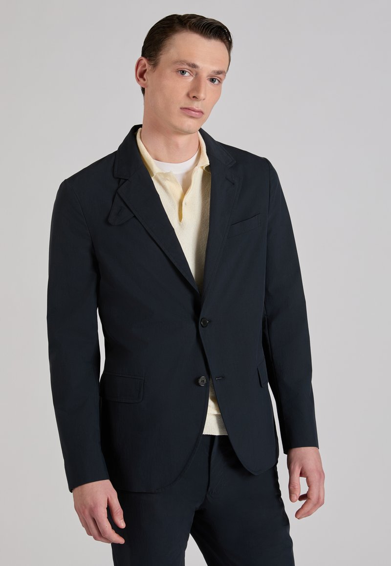 Jonge man die een zwart colbert draagt over een crèmekleurig poloshirt, staand tegen een effen lichtgrijze achtergrond.