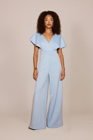 Frau mit lockigem Haar, die einen hellblauen Jumpsuit mit weitem Bein, tiefem V-Ausschnitt und flatternden Ärmeln trägt, steht vor einem beigen Hintergrund.