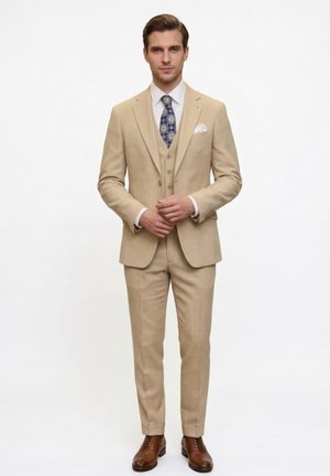 Hombre con un traje beige de tres piezas, camisa blanca, corbata azul estampada, pañuelo blanco en el bolsillo y zapatos de vestir marrones, de pie con las manos entrelazadas.