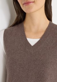Braune Strickweste mit V-Ausschnitt und strukturierter Musterung. Über einem weißen Langarmshirt getragen und sorgt für einen Lagen-Look.