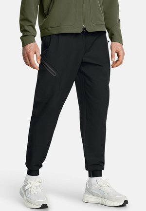 Mężczyzna ubrany w czarne spodnie dresowe z kieszenią na zamek, oliwkowozieloną kurtkę, białe skarpetki i białe trampki na jednolitym tle.