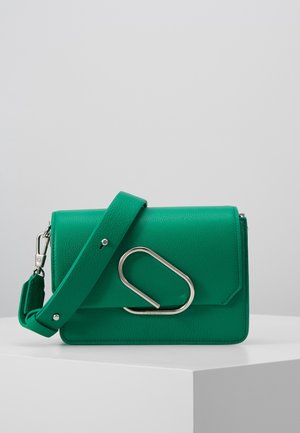 Grüne umhängetasche aus strukturiertem Leder mit breitem Riemen und großem silbernem abstraktem Metallverschluss auf weißem Podest.