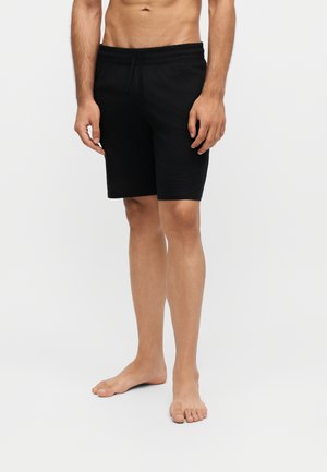 Barfod mand iført simple sorte knælange shorts stående mod en ensfarvet lys baggrund.