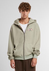 Kapturowa bluza z zamkiem błyskawicznym w jasnym zielonym kolorze, wykonana z mieszanki bawełny, z kieszenią z przodu, żebrowanymi mankietami oraz grafiką górską z napisem "ZABRONIONE".