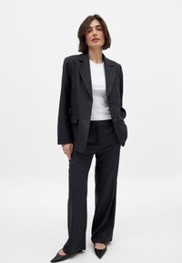 Costume à rayures fines noir avec une veste cintrée et un pantalon droit. Le tissu a une texture lisse. Porté avec un haut blanc et des talons noirs.
