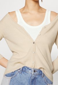 Beige Strickjacke mit tiefem V-Ausschnitt, ausgestattet mit silbernen Knöpfen und einem lässigen Schnitt, über einem weißen Tanktop getragen, mit langen Ärmeln.