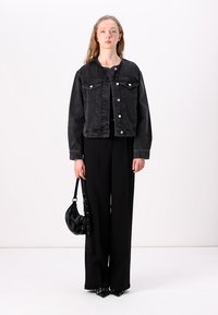 Črna jeans jakna s klasičnim dizajnom, sprednjimi žepi in srebrnimi gumbi, v kombinaciji s širokimi črnimi hlačami in majhno črno torbico.