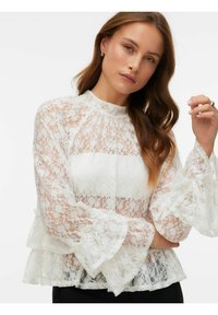 Hvit blondebluse med vide ermer og høy hals. Lagdelt design med blomsterprint og transparent tekstur, stylet over en ensfarget base.