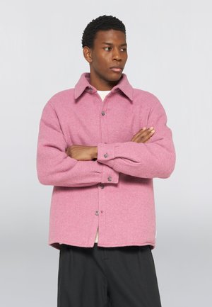 Homme aux cheveux courts et bouclés, bras croisés, portant une veste en laine rose boutonnée et un pantalon noir sur un fond uni.