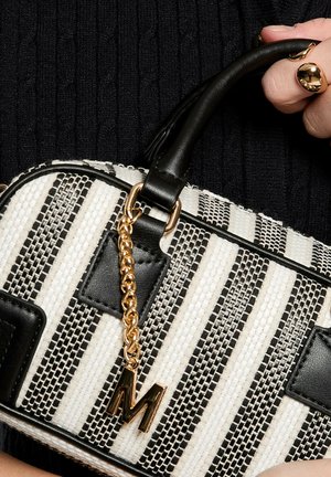 Una mano tiene una borsa intrecciata a righe bianche e nere con rifiniture in pelle nera e un ciondolo a catena dorata a forma della lettera "M".
