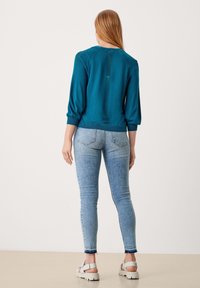 Sininen neulepaita, jossa on kolme neljänneksen hihaa ja teksturoitu takapala, yhdistetty vaaleansinisiin kapeisiin farkkuihin ja valkoisiin korollisiin sandaaleihin.