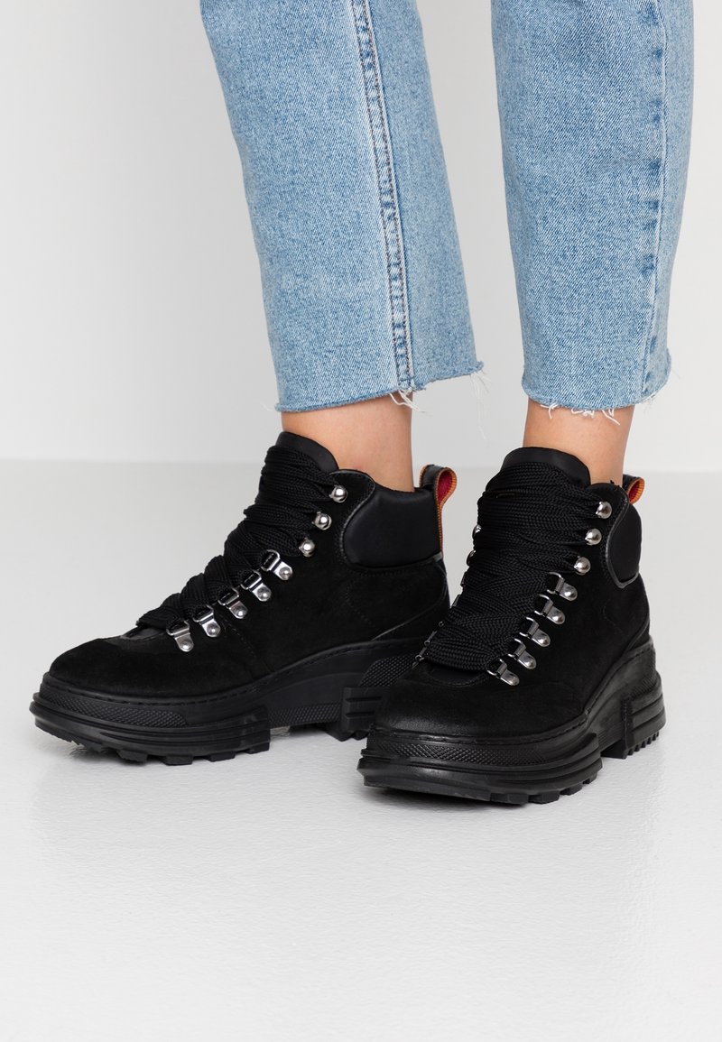 Botas altas de ante negro con suelas de goma gruesa, que cuentan con ojales metálicos, cordones gruesos negros y una lengüeta trasera en contraste de color rojo.