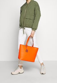 Pomarańczowa torba typu tote wykonana ze sztucznej skóry z tłoczonym wzorem, podwójnymi uchwytami i ozdobnym breloczkiem, zestawiona z zieloną pikowaną kurtką bomberką i białymi spodniami.