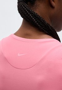 Άτομο με σκουρόχρωμο δέρμα και μακριά πλεγμένα μαλλιά που φοράει ροζ μπλούζα Nike, εμφανιζόμενο από πίσω με έμφαση στην περιοχή των ώμων και του λαιμού.