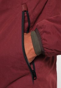 Vinröd jacka med en ficka med dragkedja, med elastiska ärmar och en slät yta. Inkluderar en kontrasterande olivgrön foderdetalj.