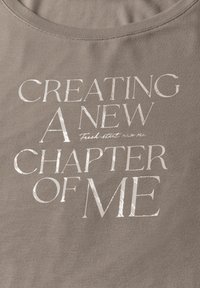 Camiseta de algodón gris con un texto metálico brillante que dice "CREANDO UN NUEVO CAPÍTULO DE MÍ" en diferentes tamaños y estilos de fuente.
