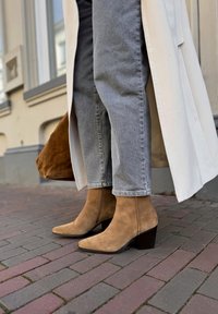 Tan suede enkellaarzen met een spitse neus en een blokhak van 5 cm, gecombineerd met lichtgrijze, iets ingekorte jeans en een lange beige jas.