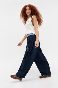 Camisola branca de malha sem mangas, cós azul acentuado, calças largas azul-marinho com bordado do logótipo e sapatos castanhos com atacadores.