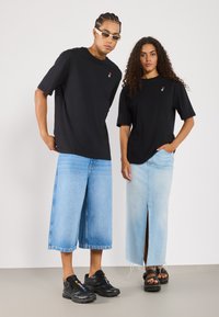 Svarte t-skjorter med liten logo, lyseblå denim culotter, og et langt denim-skjørt med splitt. Skoene inkluderer svarte joggesko og sandaler.