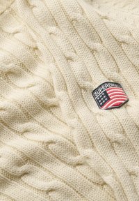 Ivory knitted sweater featuring a cable design, with a woven patch displaying the Superdry logo and an American flag.