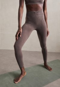 Woman standing barefoot on a green yoga mat with legs apart, wearing high-waisted brown leggings and a matching crop top.