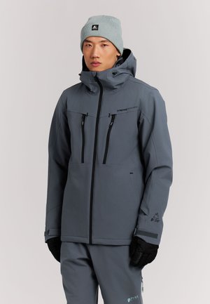 Man draagt een grijze winterjas met capuchon, bijpassende broek, zwarte handschoenen en een lichtgrijze muts tegen een effen achtergrond.