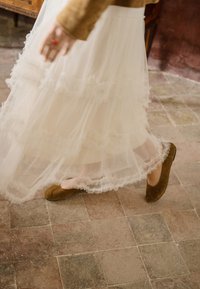 Persoană purtând o fustă stratificată din tul alb și balerini maro din piele întoarsă, mergând pe un podea cu dale, cu o piesă de mobilier din lemn în apropiere.