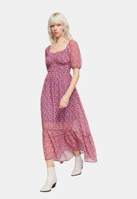 Abito maxi floreale in tonalità rosa e viola, tessuto trasparente, maniche a sbuffo, corpino aderente, gonna a strati, abbinato a stivali bianchi alla caviglia.