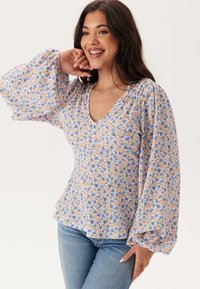 Blouse florale avec un décolleté en V, des manches longues bouffantes et un fond bleu clair orné de fleurs orange et violettes. Tissu doux et léger.