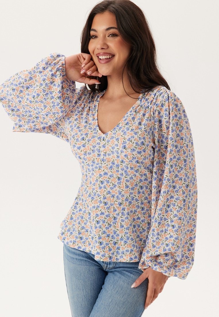 Blouse florale avec un décolleté en V, des manches longues bouffantes et un fond bleu clair orné de fleurs orange et violettes. Tissu doux et léger.