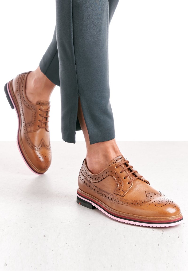 Chaussures brogue en cuir marron avec des semelles soulignées de rose, portées avec un pantalon court vert foncé dévoilant une fente à la cheville.