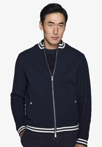 Marineblå zip-up jakke med kabelstrikkede ærmer, ribbet krave og håndled, begge med hvide striber, samt to sidelommer.
