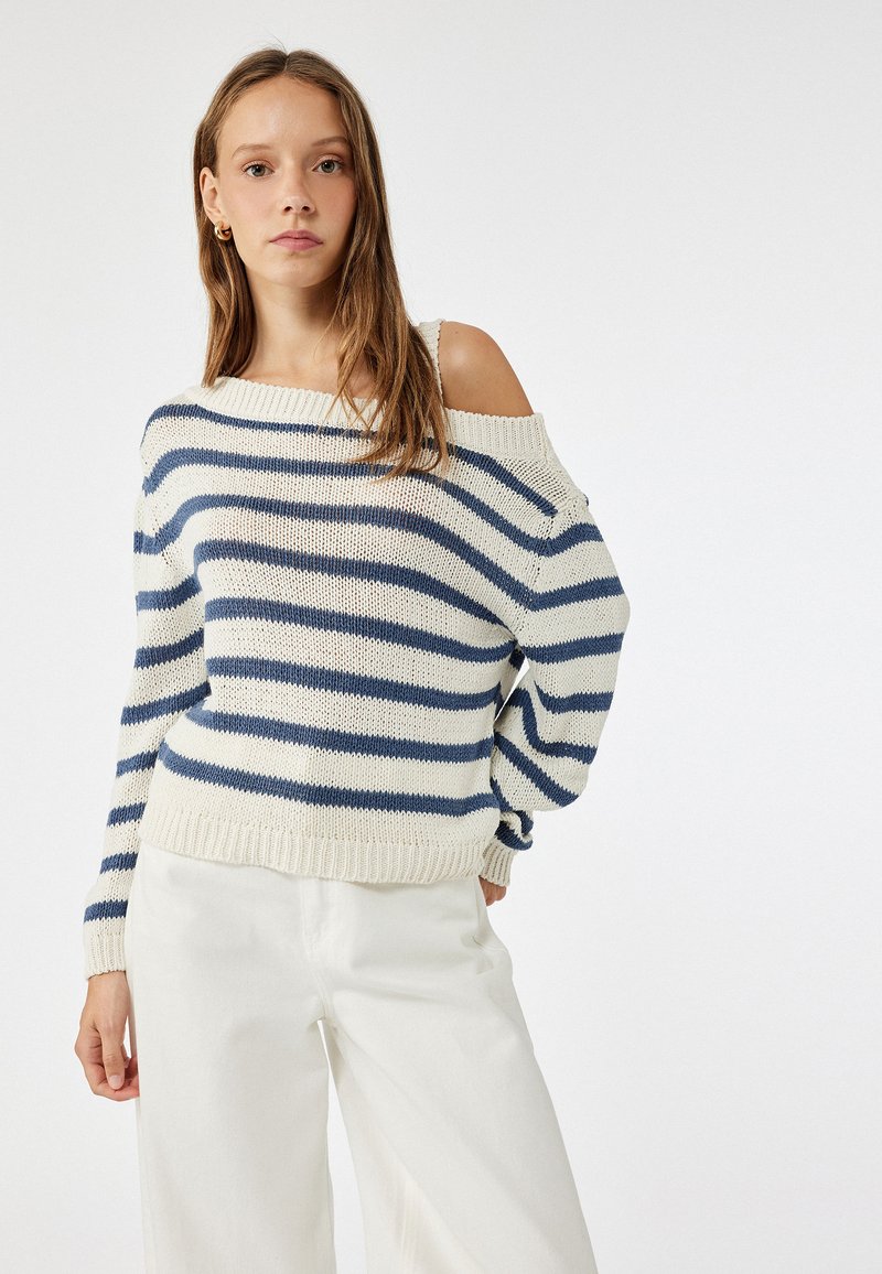 Suéter de punto a rayas en crema y azul marino, con un diseño off-shoulder, puños acanalados y un corte holgado. Combinado con pantalones anchos blancos.