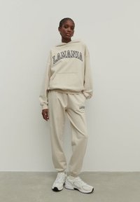 Bežinė hoodie ir treningo kelnės su tamsiai mėlyna "LAMANIA" užrašą, su kengūros kišene ir elastingomis rankogaliais. Dėvimos su baltais sportiniais batais.
