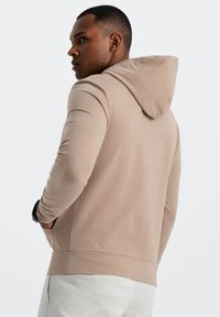 Felpa beige con una vestibilità rilassata e cappuccio regolabile, dotata di maniche lunghe e una texture liscia. Senza motivi visibili o accenti.