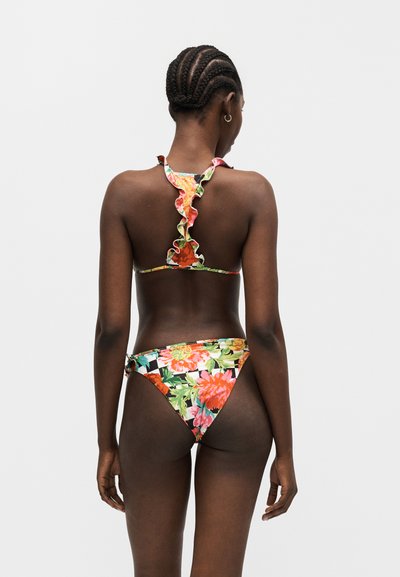 Γυναίκα με πλεξούδες που φοράει φλοράλ μπικίνι με βολάν, εμφανίζοντας την πίσω όψη σε μονόχρωμο ανοιχτό γκρίζο φόντο.