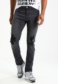 Slim-fit svarta denimjeans med slitna detaljer och rivna knän. Dessa är kombinerade med grå sportskor och en vit t-shirt med tryck.