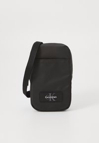 Sac en bandoulière en nylon noir de forme rectangulaire, avec une sangle et un patch logo à l'avant. Texture lisse, design minimaliste et fermeture zippée.