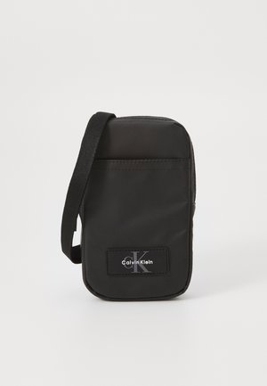 Sac en bandoulière en nylon noir de forme rectangulaire, avec une sangle et un patch logo à l'avant. Texture lisse, design minimaliste et fermeture zippée.