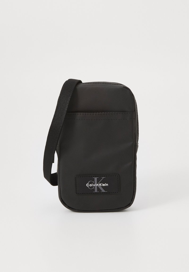 Sac en bandoulière en nylon noir de forme rectangulaire, avec une sangle et un patch logo à l'avant. Texture lisse, design minimaliste et fermeture zippée.