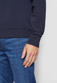 Pullover blu navy con polsini a costine, abbinato a jeans blu. Il tessuto appare liscio e la vestibilità è rilassata attorno al torso e alle braccia.
