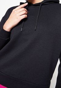 Personne portant un sweat à capuche noir uni avec cordons, main touchant le cou, cadrée du menton à la taille, fond neutre.