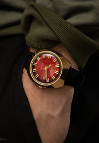 Rote Ziffernblattuhr mit Gehäuse in Goldfarbe, schwarzem Armband und auffälligen Stundenmarkierungen. Verfügt über ein Quarzwerk und eine Datumsanzeige auf der Position 6 Uhr.