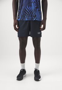 Pantalones cortos deportivos en color navy con un logotipo bordado de EA7. Combinados con una camiseta azul con estampado y zapatillas negras con detalles en blanco.